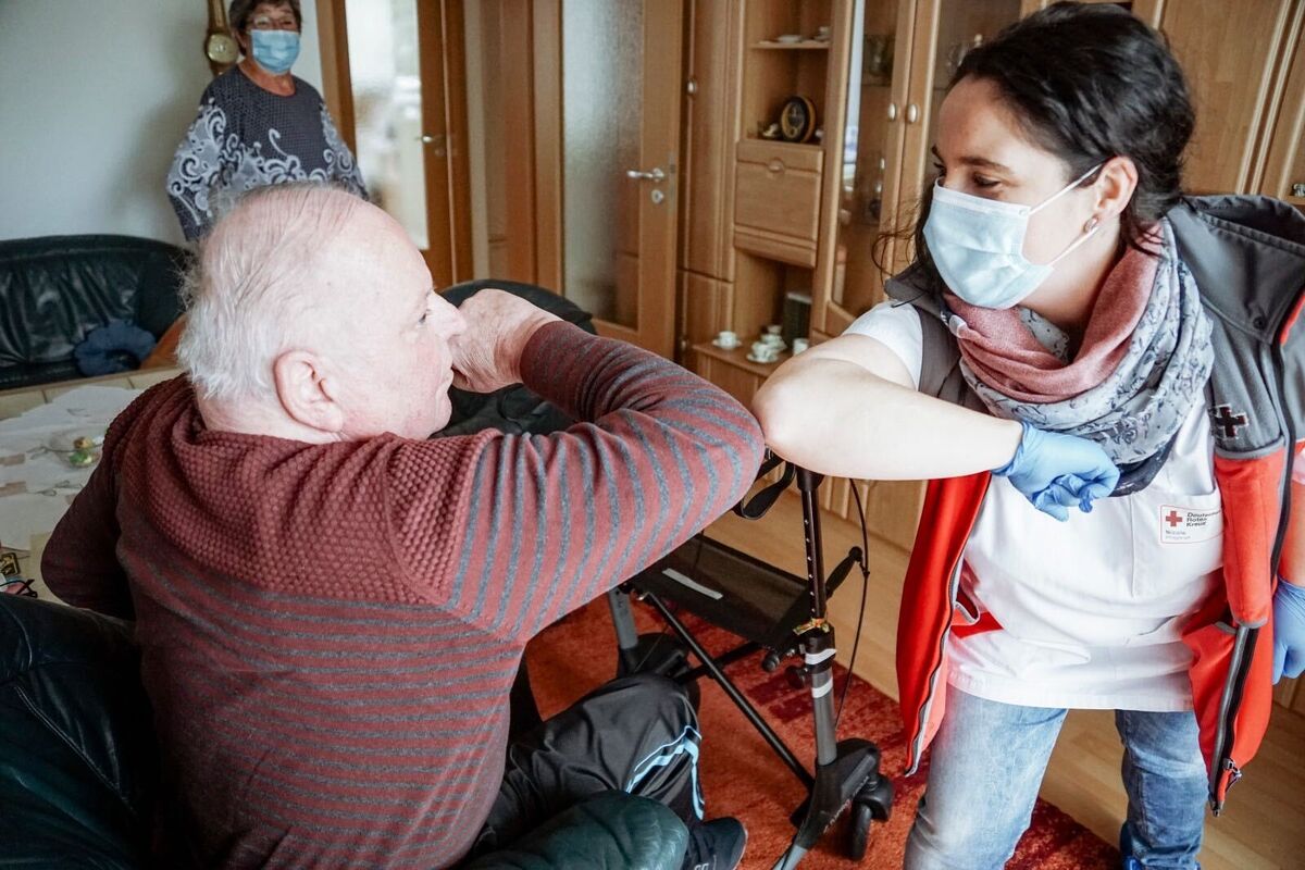 Eine Pflegekraft mit Mundschutz und ein älterer Mann berühren sich zur Begrüßung Ellenbogen an Ellenbogen.